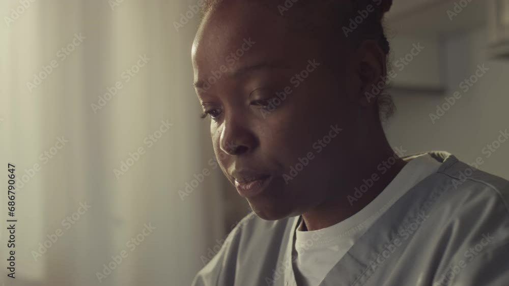 Closeup tilting shot of young African American woman in medical uniform sitting at table in kitchen in morning, eating sandwich, drinking tea or coffee and typing message on smartphone before shift Closeup tilting shot of young African American woman in medical uniform sitting at table in kitchen in morning, eating sandwich, drinking tea or coffee and typing message on smartphone before shift