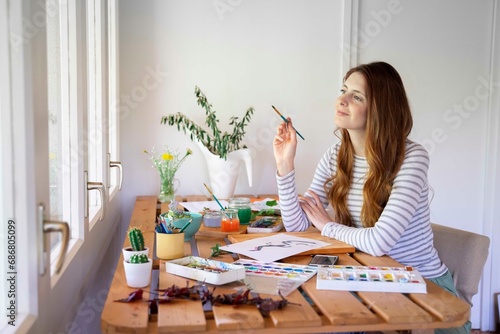 Thoughtful young woman holding paintbrush while painting on table at home