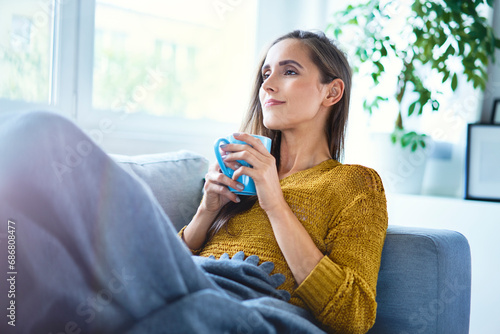 Beautiful young woman enjoying cup of coffee while lying on sofa under blanket