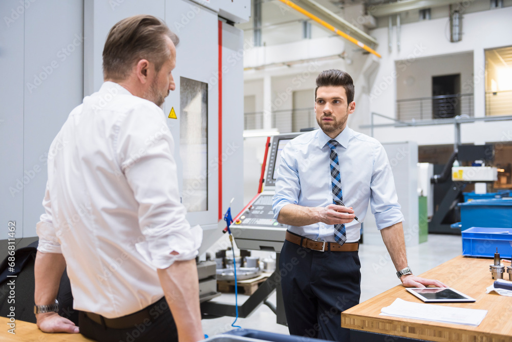 Two businessmen at table in factory shop floor discussing Two businessmen at table in factory shop floor discussing