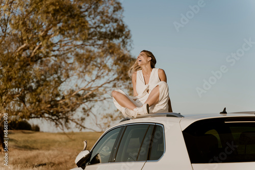 Laughing female traveller sitting on white car roof