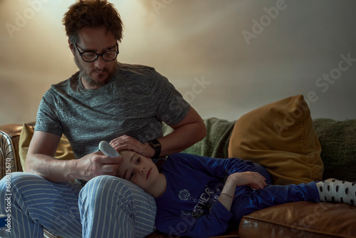 Man holding thermometer on sick daughter's forehead in living room at home photo