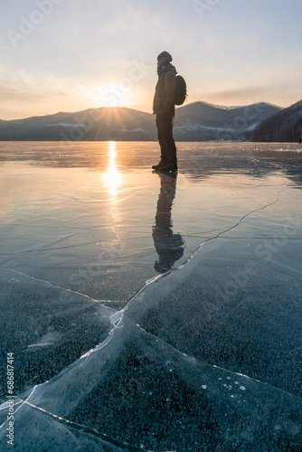 A hiker in winter clothes stands on a frozen ice surface. The mirror-smooth surface of lake ice and a person standing on it during sunset.