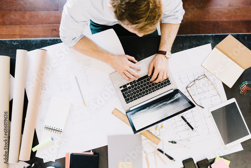 Man using laptop next to construction plan at desk, top view