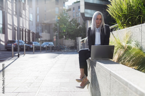 Young businesswoman sitting on a wall in the city using laptop photo