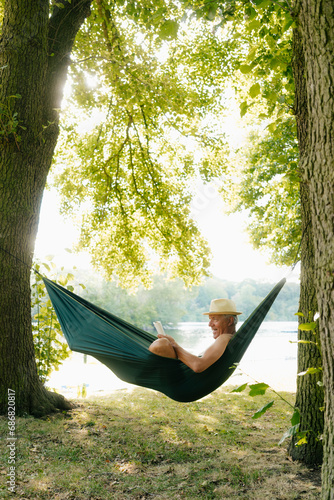 Senior man wearing straw hat relaxing in hammock at lakeshore reading book