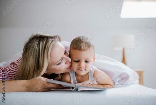 Mother kissing toddler son lying in bed at home reading a book