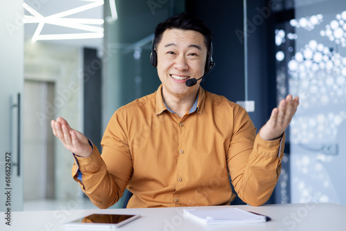 Fototapeta Portrait of a smiling young asian man sitting in the office at a table in front of a camera in a headset, spreading his hands, smiling at the camera