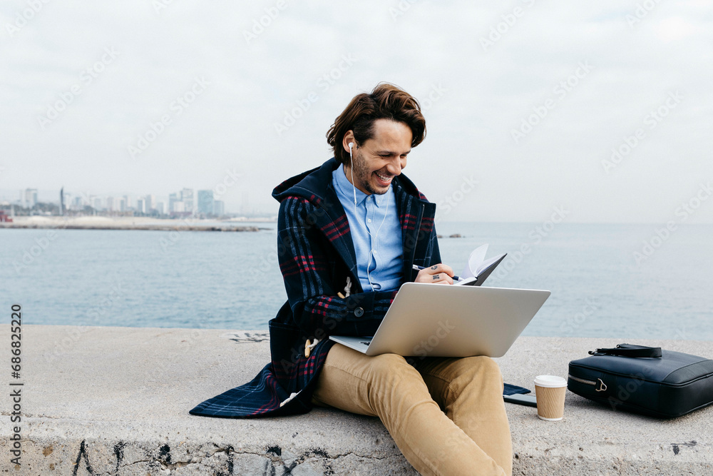 Spain, Barcelona, smiling man sitting at the sea working with laptop and notebook