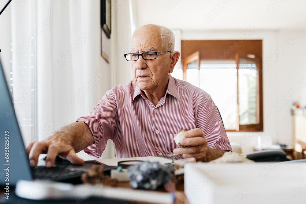 Retired elderly man using laptop while doing research on fossils and minerals at home Retired elderly man using laptop while doing research on fossils and minerals at home