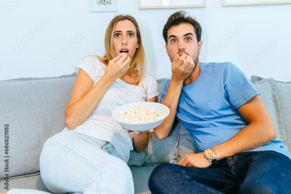 Excited couple watching TV while eating popcorn on sofa at home