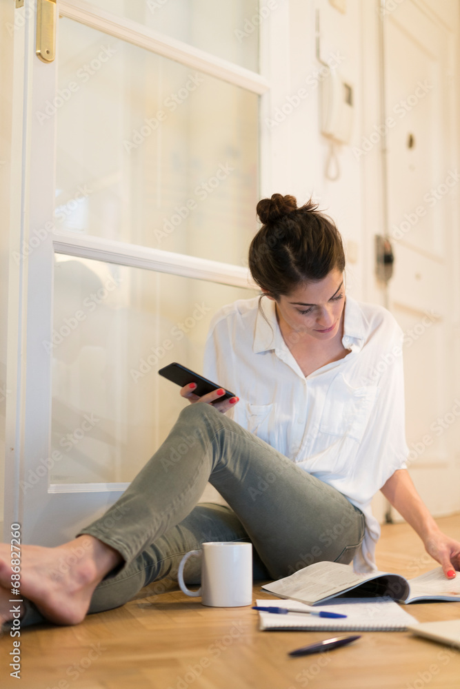 Young woman sitting on the floor with coffee mug and smartphone looking at booklet Young woman sitting on the floor with coffee mug and smartphone looking at booklet