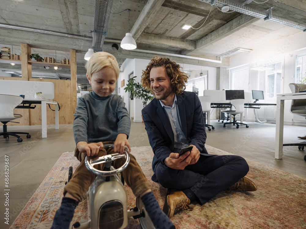 Smiling businessman with son on toy car in office