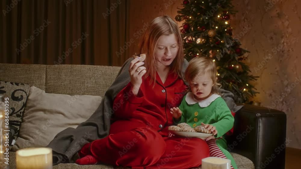 Mother with a toddler sitting on a couch and eating gingerbread cookies, having holiday time together. Woman in red pajamas and Christmas tree in the background plays with a child. Mother with a toddler sitting on a couch and eating gingerbread cookies, having holiday time together. Woman in red pajamas and Christmas tree in the background plays with a child.