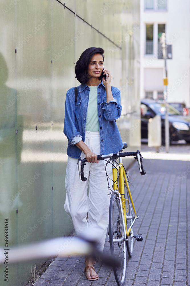 Female commuter talking over smart phone while standing with bicycle on street in city Female commuter talking over smart phone while standing with bicycle on street in city