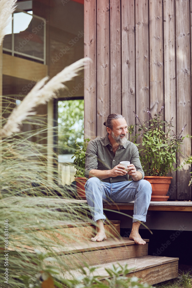 Thoughtful mature man holding coffee cup while sitting outside tiny house