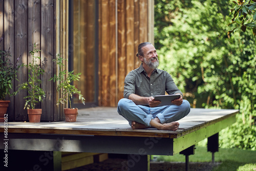 Bearded mature man holding digital tablet contemplating while sitting outside tiny house