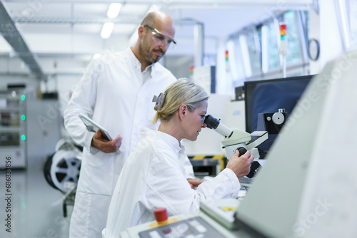 Male technician standing by female scientist looking into microscope while doing research in laboratory photo