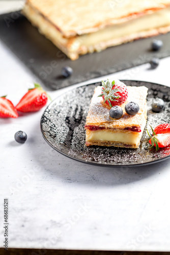 A piece of Napoleon cake with poppy seeds and cherries, decorated with strawberries, blueberries on a light background. Close up