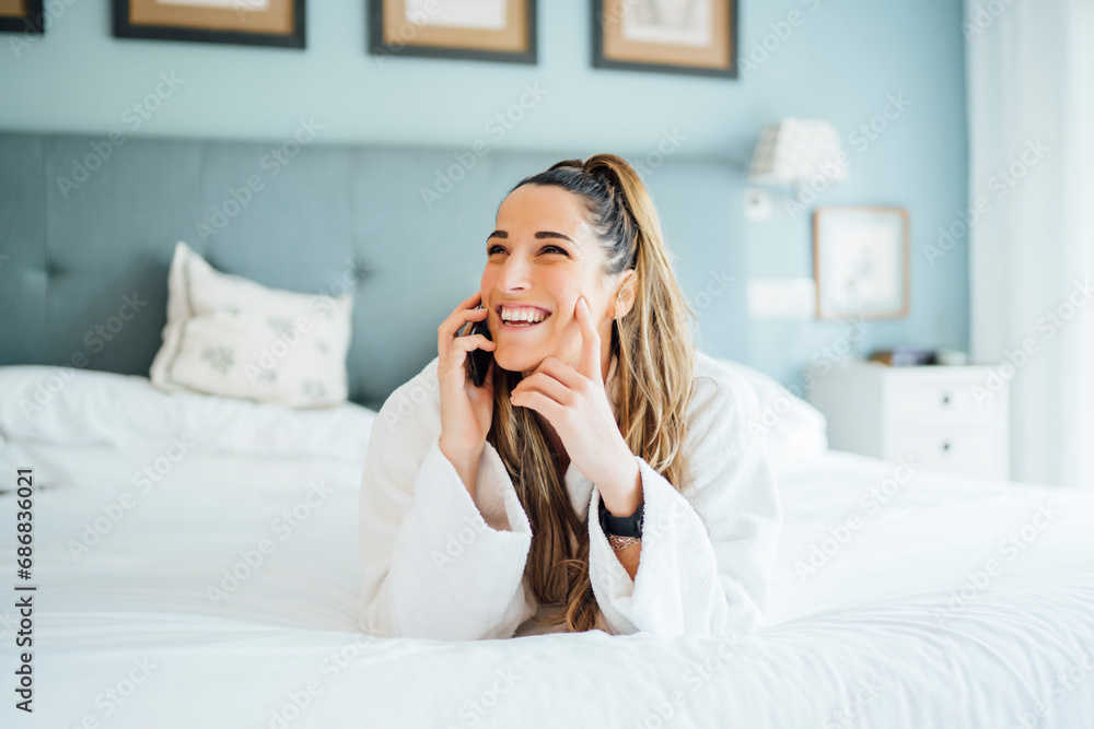 Cheerful mid adult woman talking over smart phone while lying on bed in hotel Cheerful mid adult woman talking over smart phone while lying on bed in hotel