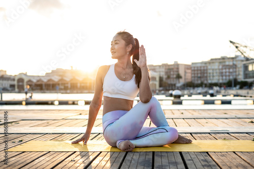 Asian woman practicing yoga on a pier at harbour, half-spinal twist at sunset