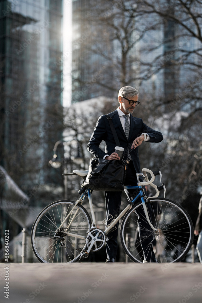 Grey-haired businessman with bicycle and coffee to go in the city looking at smartwatch