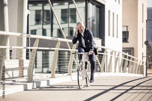 Smiling mature businessman with headphones riding bicycle on a bridge in the city