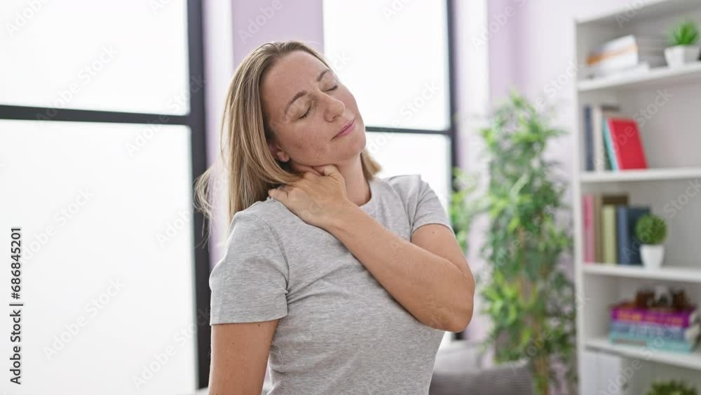 Worried young blonde woman suffering severe neck pain at home, standing alone in the living room, resting her achy cervical amidst cozy interior Worried young blonde woman suffering severe neck pain at home, standing alone in the living room, resting her achy cervical amidst cozy interior