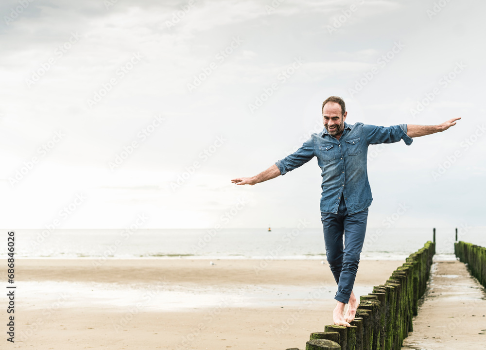 Carefree man with arms outstretched walking on wooden post at beach Carefree man with arms outstretched walking on wooden post at beach