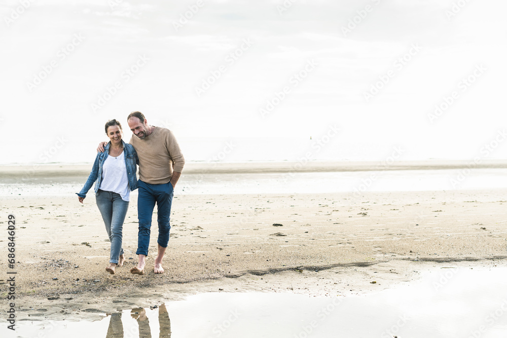 Smiling mature couple walking at beach against sky during sunset