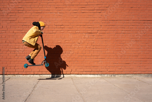 Boy performing stunt with push scooter on sidewalk against brick wall during sunny day