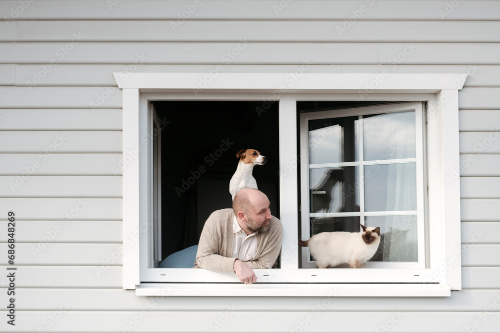 Mature man with cat and dog leaning out of window of his house