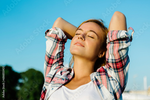 Woman with eyes closed hands behind head standing against clear sky