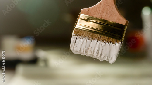 The close up shot shows a paintbrush a construction brush with white paint on the pile. It is used to cover the surface in repair work. The background is blurred in the frame it is hanging from above
