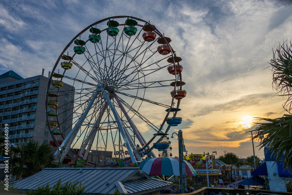 A beautiful summer landscape at the Carolina Beach Boardwalk with Ferris wheels and carnival rides, people, palm trees and grass, blue sky and clouds at sunset in Carolina Beach North Carolina USA