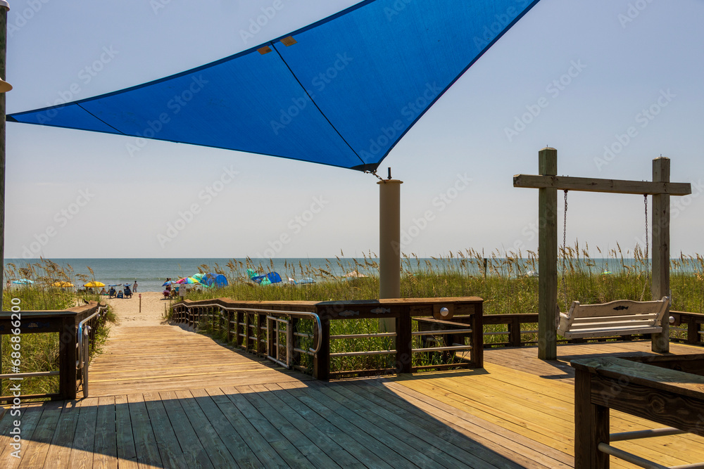 A beautiful summer landscape at the beach with a wooden footpath and swing, people relaxing on the beach with colorful umbrellas, blue ocean water and blue sky in Carolina Beach North Carolina USA