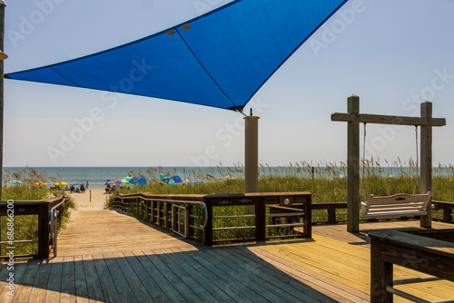 A beautiful summer landscape at the beach with a wooden footpath and swing, people relaxing on the beach with colorful umbrellas, blue ocean water and blue sky in Carolina Beach North Carolina USA