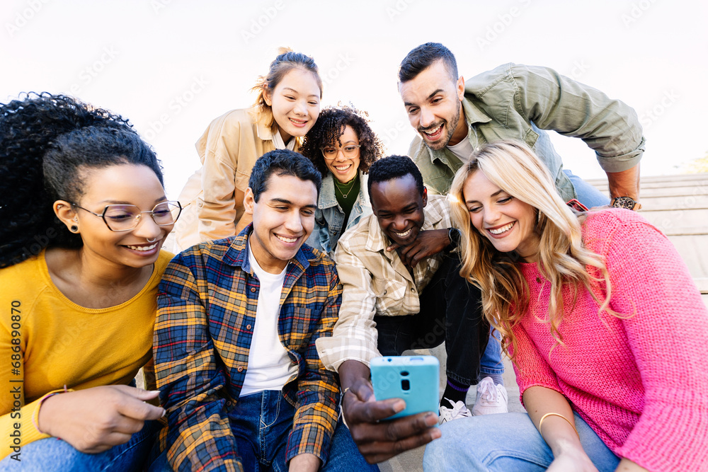 Young group of diverse friends looking at mobile phone screen. Millennial people laughing while watching social media content on cell smartphone feed app sitting together outdoors. Young group of diverse friends looking at mobile phone screen. Millennial people laughing while watching social media content on cell smartphone feed app sitting together outdoors.