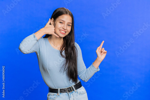 Call me, here is contact number. Caucasian young woman looking at camera doing phone gesture asking for conversation. Hotline online service proposition advertisement. Girl isolated on blue background
