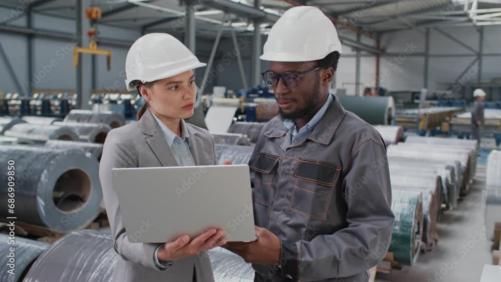 Handsome foreman with working uniform and safety helmet explaining something to beautiful manager. Young lady holding laptop and actively listening to worker. Industrial manufacturing. Workshop. Handsome foreman with working uniform and safety helmet explaining something to beautiful manager. Young lady holding laptop and actively listening to worker. Industrial manufacturing. Workshop.