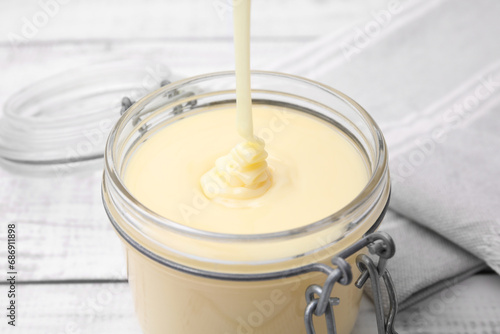 Pouring tasty condensed milk into jar on white table, closeup photo