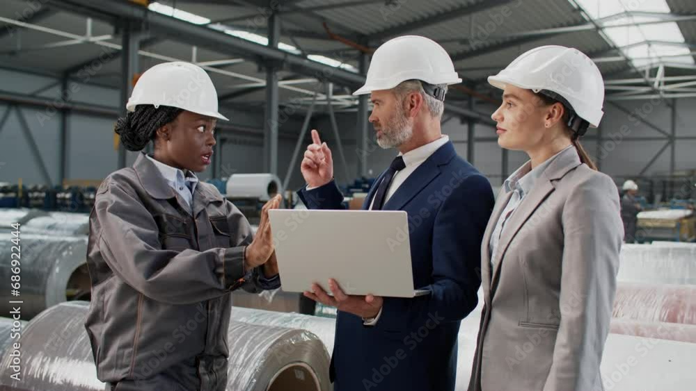 Hardworking forewoman talking with owners factory inside. Multiethnic people with helmets having conversation in warehouse. Attractive businessman holding laptop while speaking with female worker. Hardworking forewoman talking with owners factory inside. Multiethnic people with helmets having conversation in warehouse. Attractive businessman holding laptop while speaking with female worker.