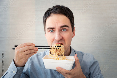 young man in a blue shirt is eating noodles from a box with a dissatisfied face. Lunch at the office. tasteless junk food photo
