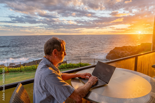 Mature Caucasian man sitting at a round table on a balcony overlooking the ocean working on a laptop watching the sun setting into the horizon, Poipu, Kauai, Hawaii