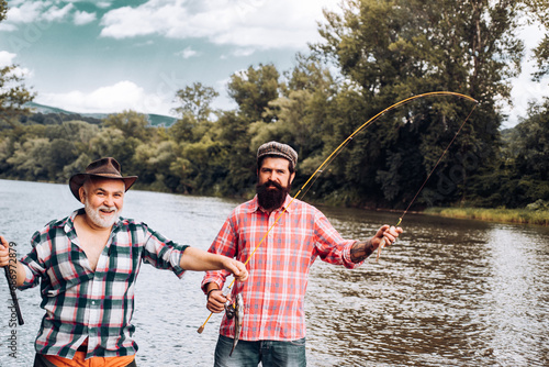 Two men friends fishing. Flyfishing angler makes cast, standing in river water. Old and young fisherman.