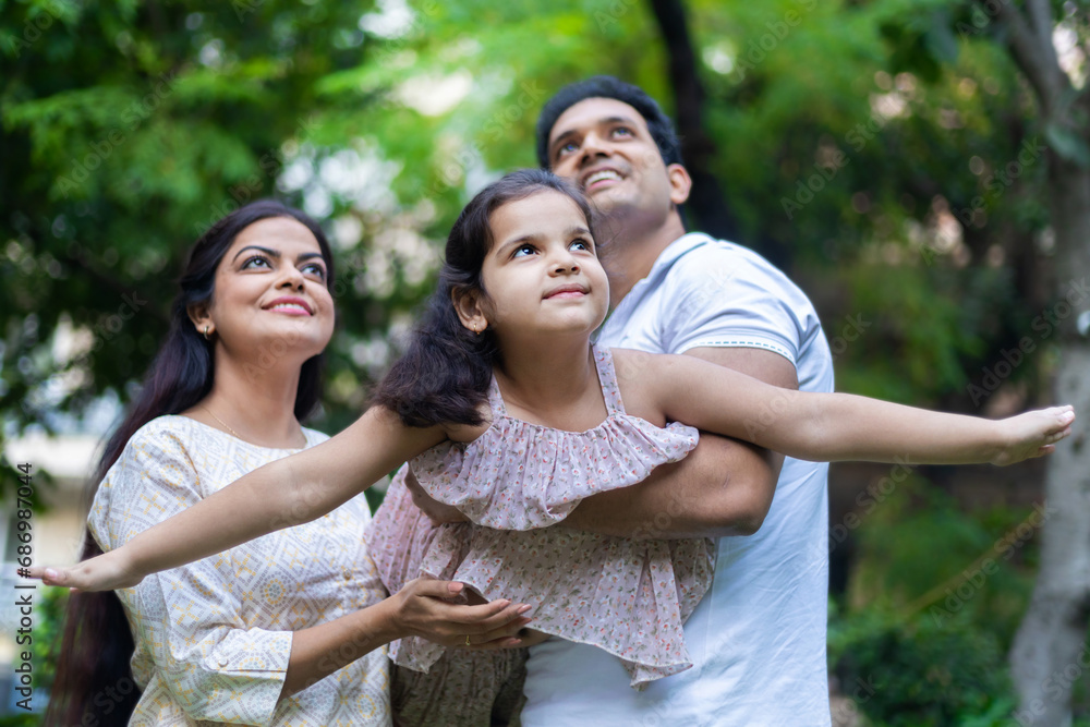 Happy indian family of three father holding daughter in arms playing flying plane, mother looking up enjoying in garden or park. Child dream concept