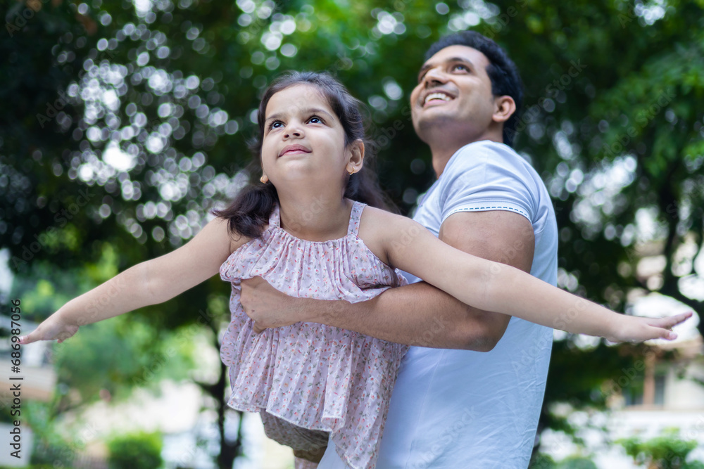 Happy indian father holding daughter in arms playing flying plane, enjoying leisure time together in garden or park. Child dream concept Happy indian father holding daughter in arms playing flying plane, enjoying leisure time together in garden or park. Child dream concept