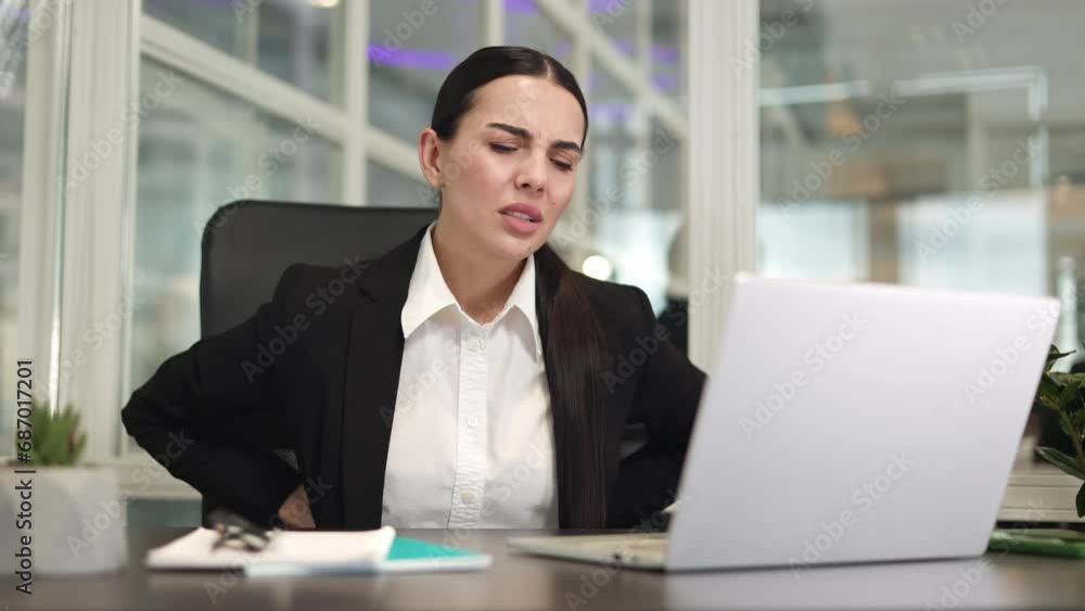 Exhausted business woman sitting at table and working with digital laptop while feeling severe back pain. Adult caucasian employee wearing black jacket trying stretching back on office chair indoor. Exhausted business woman sitting at table and working with digital laptop while feeling severe back pain. Adult caucasian employee wearing black jacket trying stretching back on office chair indoor.