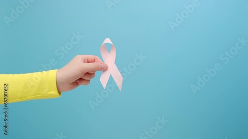 Closeup of woman hand holding small pink ribbon, symbol of breast cancer awareness, oncological disease prevention and female healthcare, posing isolated over blue color background wall in studio photo
