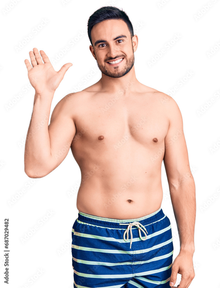 Young handsome man wearing swimwear waiving saying hello happy and smiling, friendly welcome gesture Young handsome man wearing swimwear waiving saying hello happy and smiling, friendly welcome gesture